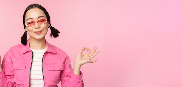 Mindfulness and wellness concept. Smiling korean girl in stylish outfit meditating, listening mantra, holds hands in zen, peace pose, practice yoga, standing over pink background