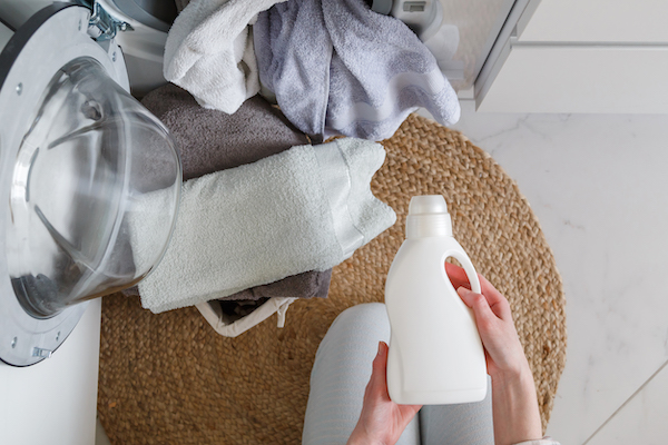 Woman holds bottle with liquid laundry detergent before washing clothes. Washer and clothes ready for laundry. View from above