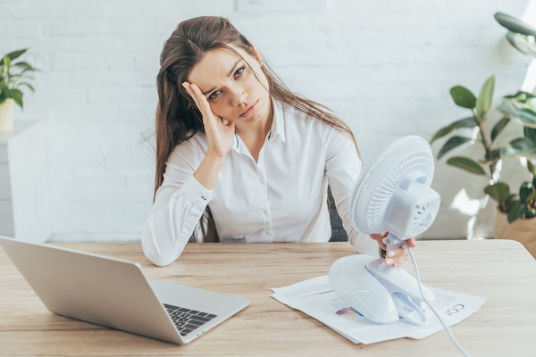 upset businesswoman sitting at workplace with paperwork, laptop and electric fan