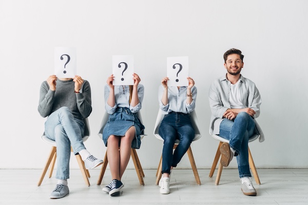 Handsome man smiling at camera near employees holding cards with question marks while waiting job