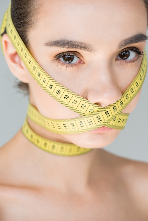 portrait of attractive young woman with closed mouth by measurement tape isolated on gray background