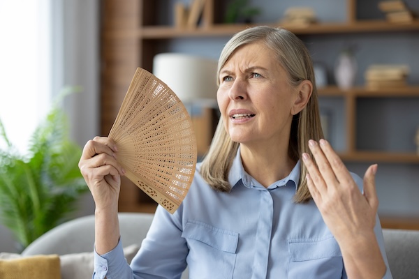 Disturbed woman waving with wooden hand fan to squinted face while sitting in overheated apartment. Exhausted woman suffering from no air conditioner at home during summer temperature increasing