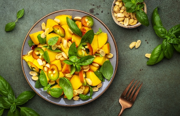 Exotic gourmet salad with juicy sweet peach, hot jalapeno peppers, salted peanuts and green basil, rustic green table background, top view