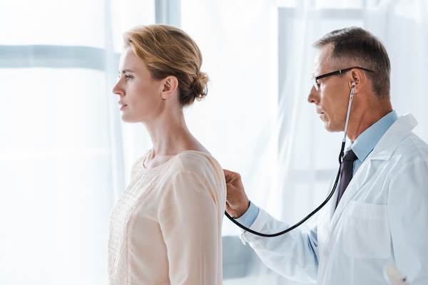 side view of doctor in glasses using stethoscope while examining attractive patient in clinic
